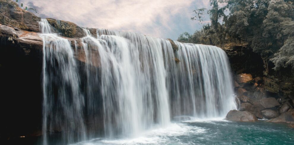Peaceful waterfall cascading over rocks with lush forest backdrop and serene sky.